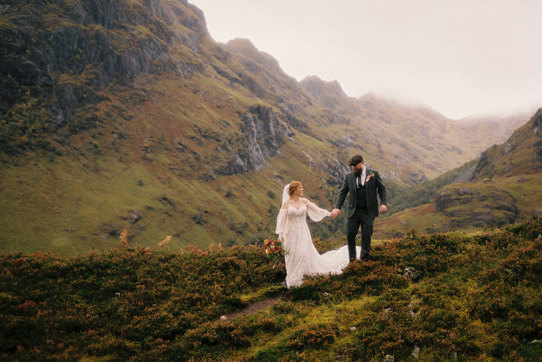 bride and groom hold hands as they walk through the scottish hills on wedding day