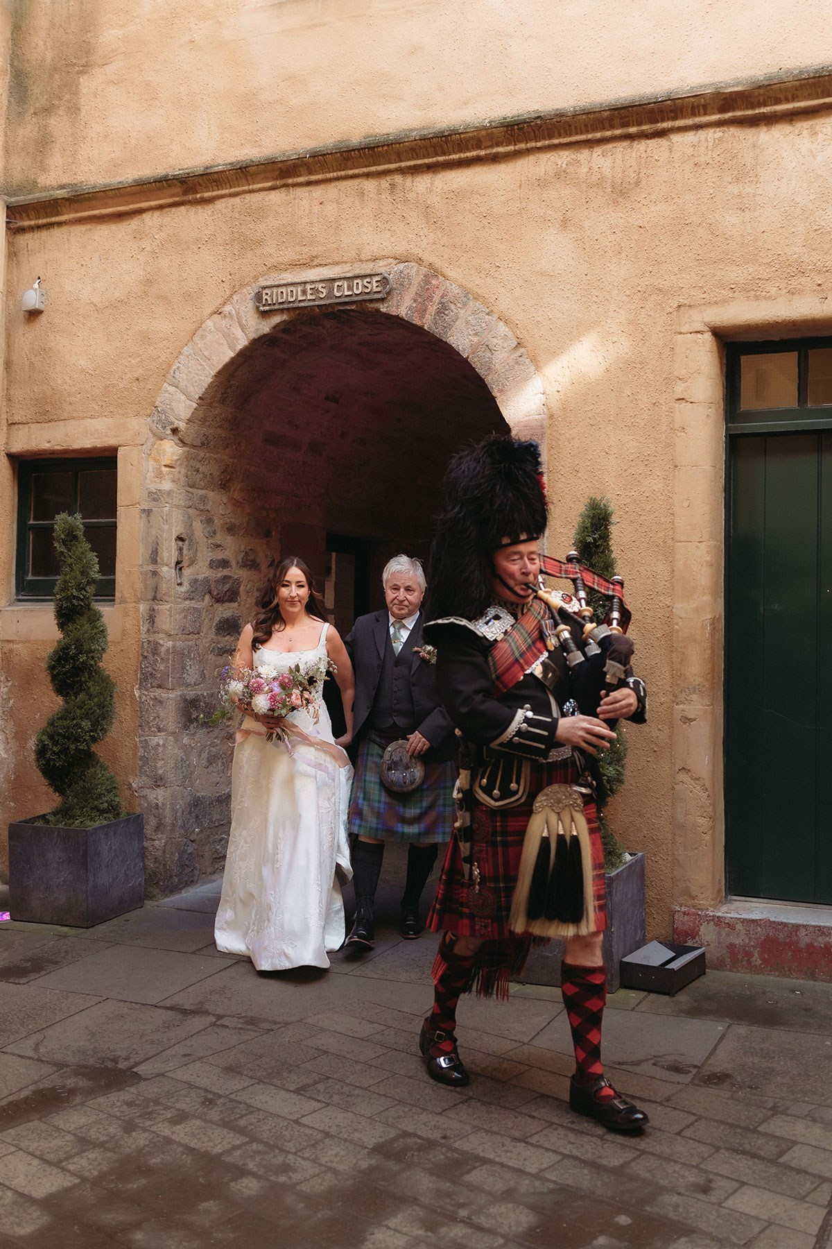 a man in a kilt playing the bagpipes walking in front of a bride and a man at Riddle's Court in Edinburgh
