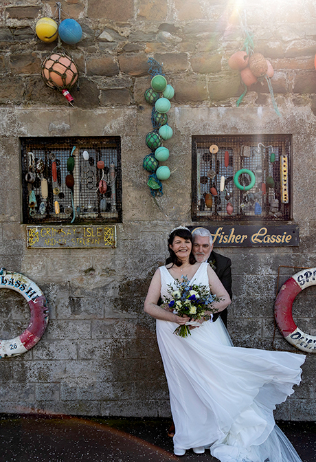 a windswept bride and groom standing in front of a stone building that's adorned with sea paraphernalia