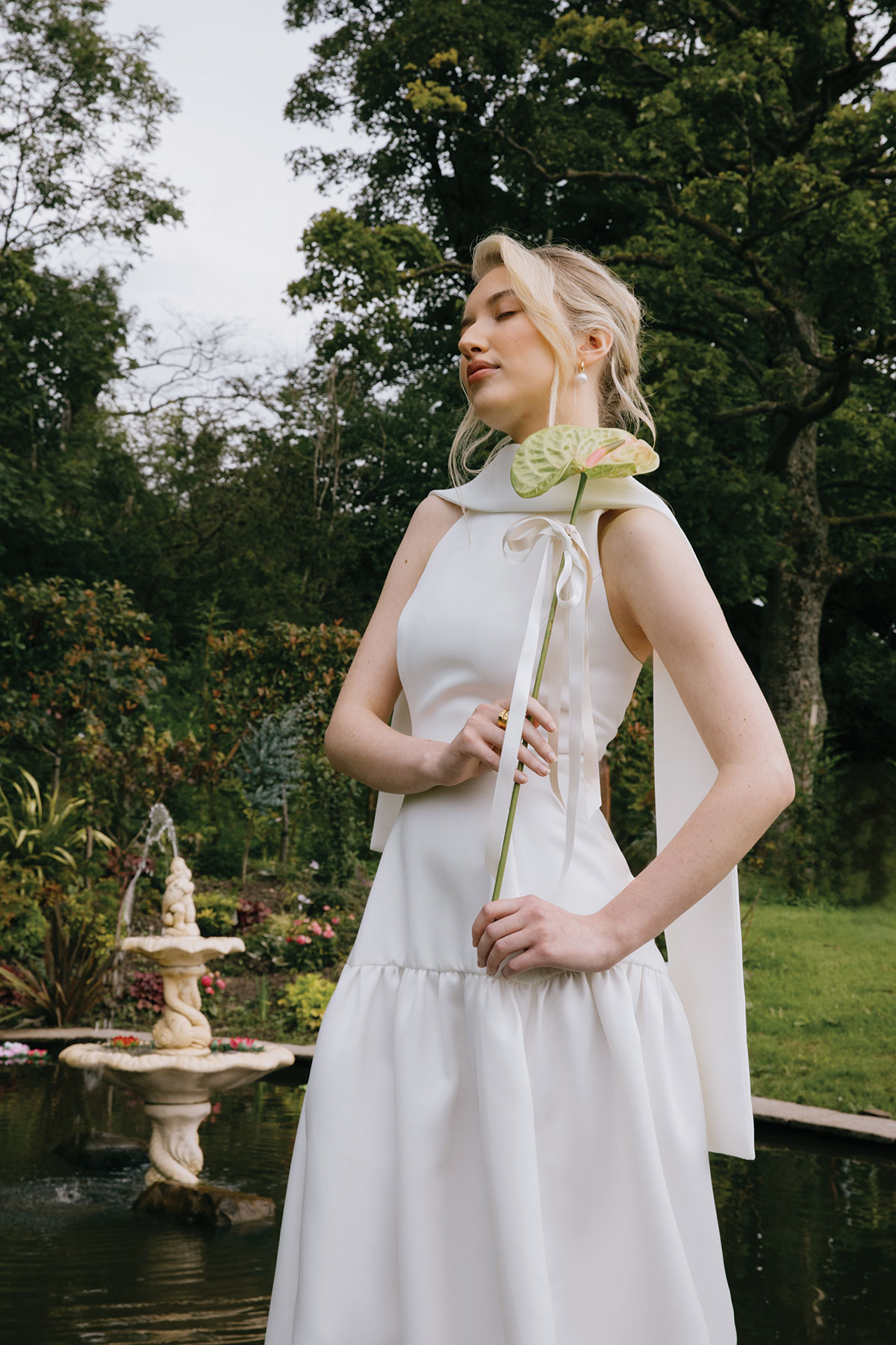 A bride in a white gown holds a single green anthurium flower while walking through a landscaped garden with fountain.