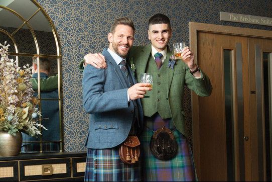 Two men wearing kilts hold up a glass of whisky to the camera with their arms around each other