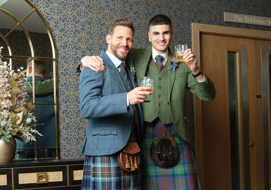 Two men wearing kilts hold up a glass of whisky to the camera with their arms around each other