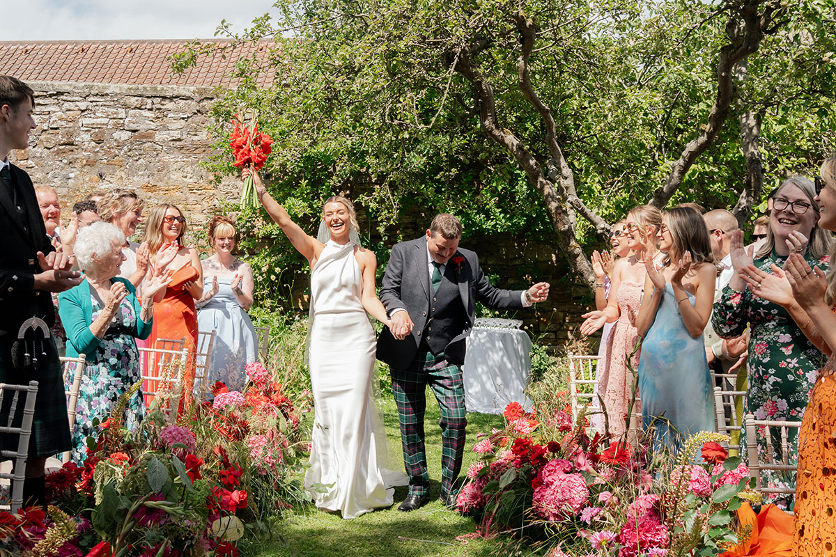 Bride and her father celebrate as they walk down the flower-lined aisle following an outdoor wedding ceremony at Kinkell Byre in Fife.