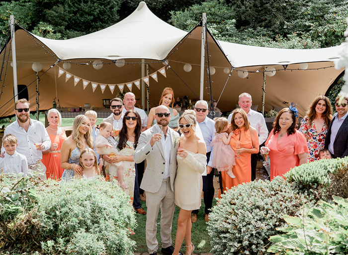 a bride, groom and group of wedding guests line up outside a canvas stretch tent in a garden