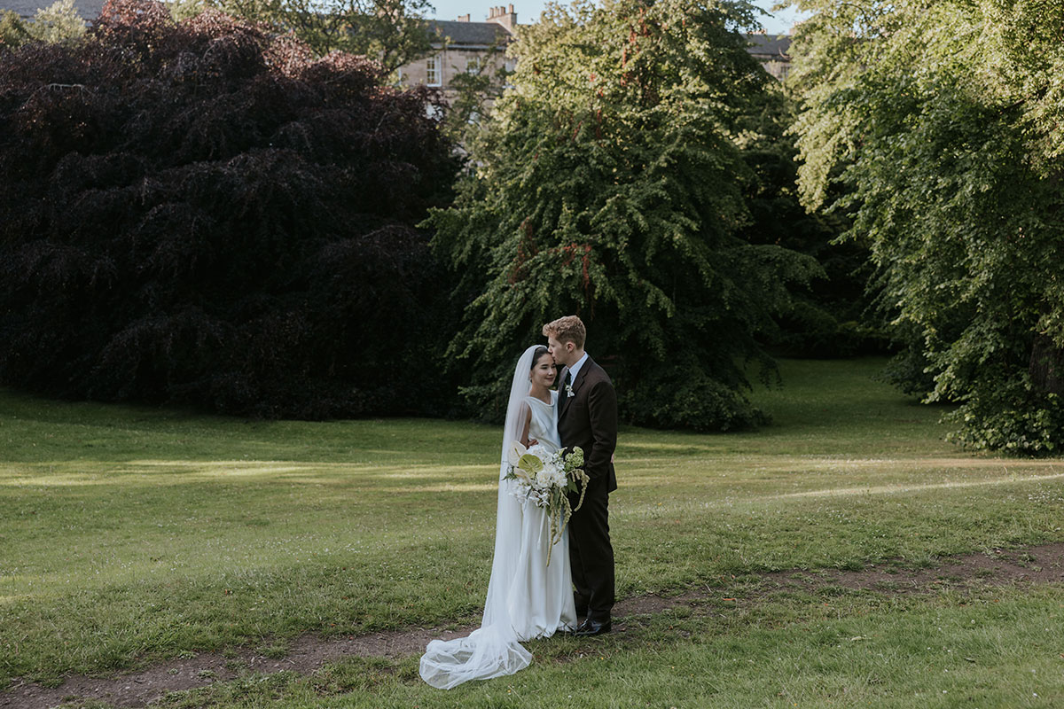 Bride and groom embracing on a grassy lawn surrounded by tall trees.