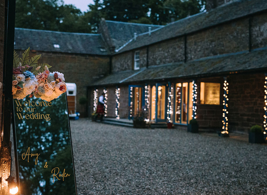 A stone building covered in fairylights with gravel ground in the background and a wedding sign covered in flowers that reads 'welcome to our wedding Amy & Roden