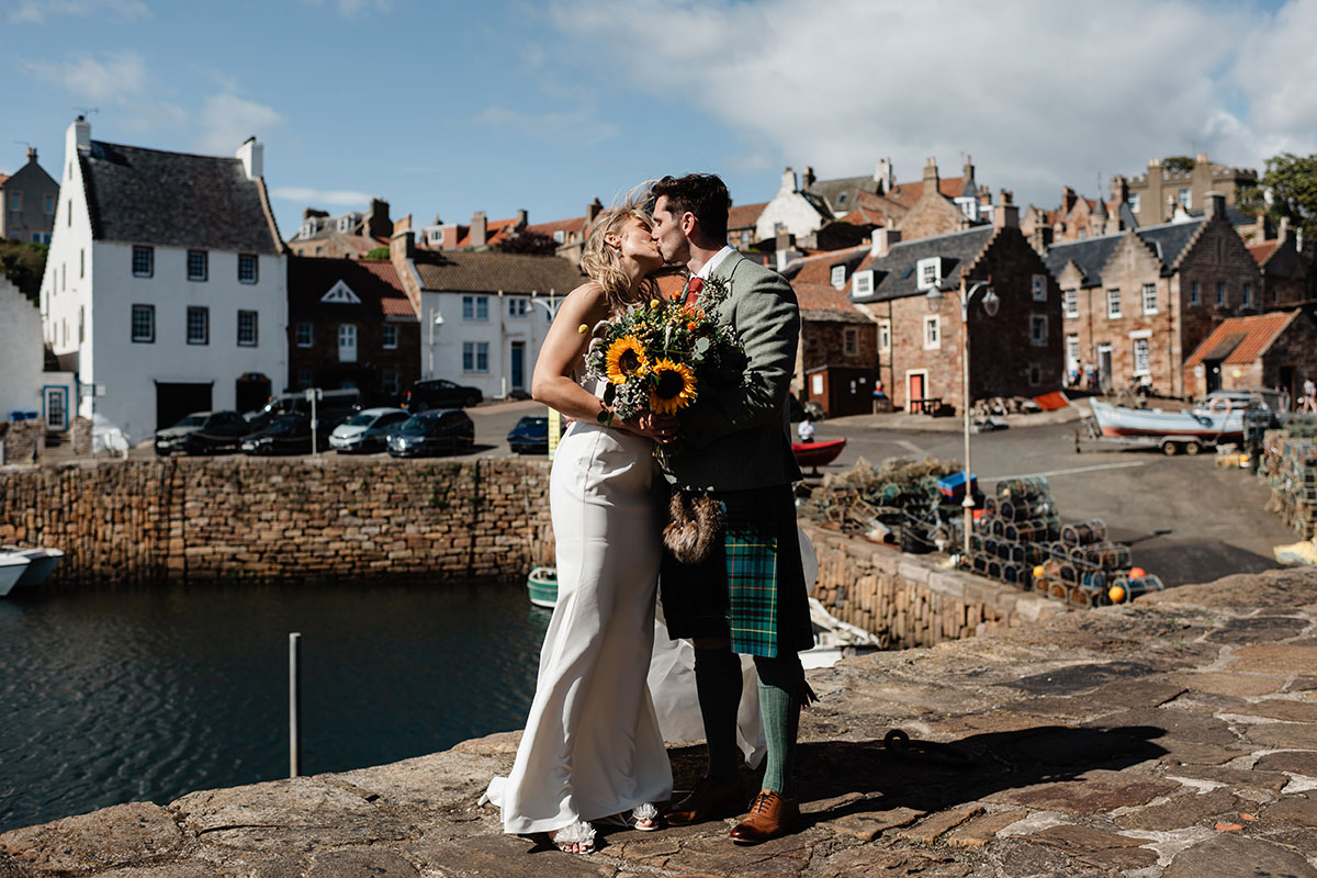 Bride and groom kissing beside the harbour wall in a coastal village, holding a sunflower bouquet