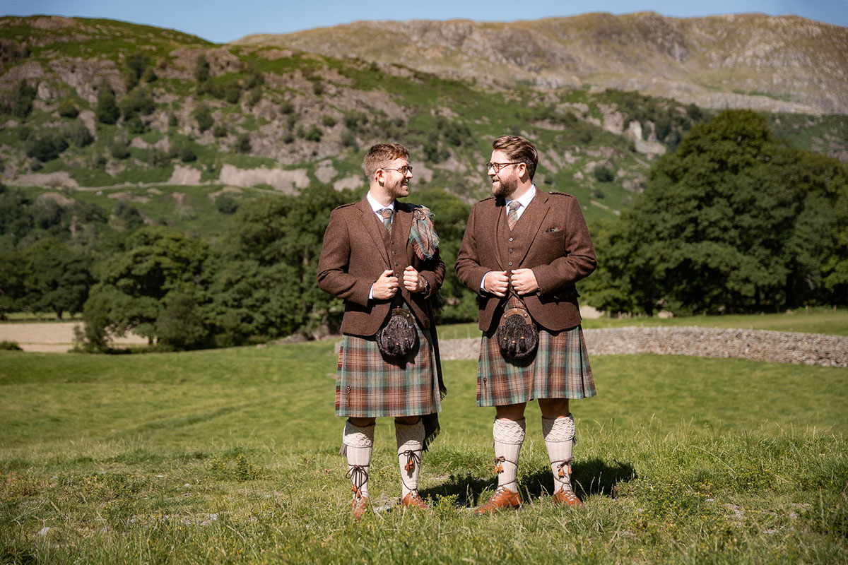 Groom and best man stand outdoors in a grassy field with hills behind them. Both wear brown tweed jackets with tartan kilts and sporrans, smiling at each other in the sunshine