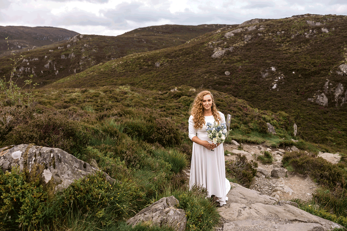 Bride standing on a rocky mountain path holding a bouquet during a Scottish elopement ceremony, surrounded by rolling Highland hills and heather-covered landscape