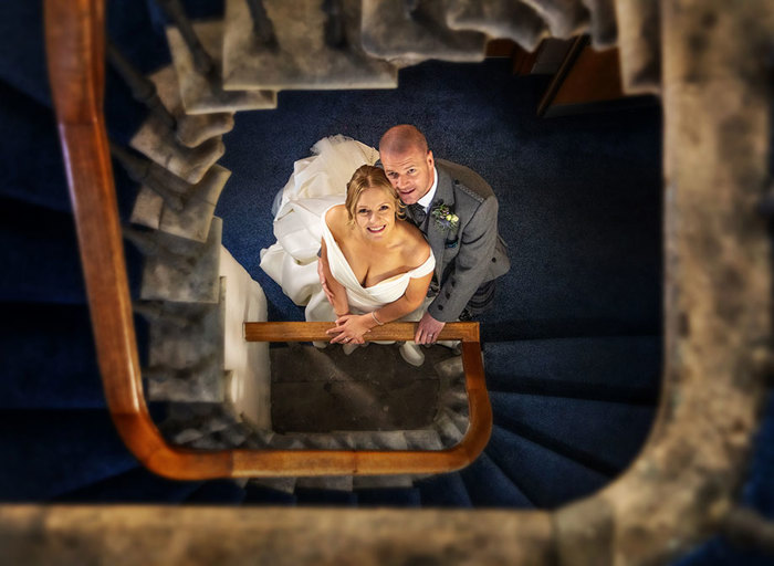 a bride and groom stand in a carpeted stairwell looking straight up at the camera