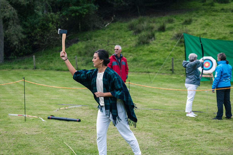 A Person Standing On Grass Wearing A Tartan Shawl Holding An Axe Aloft While Two Other People Do Archery In Background