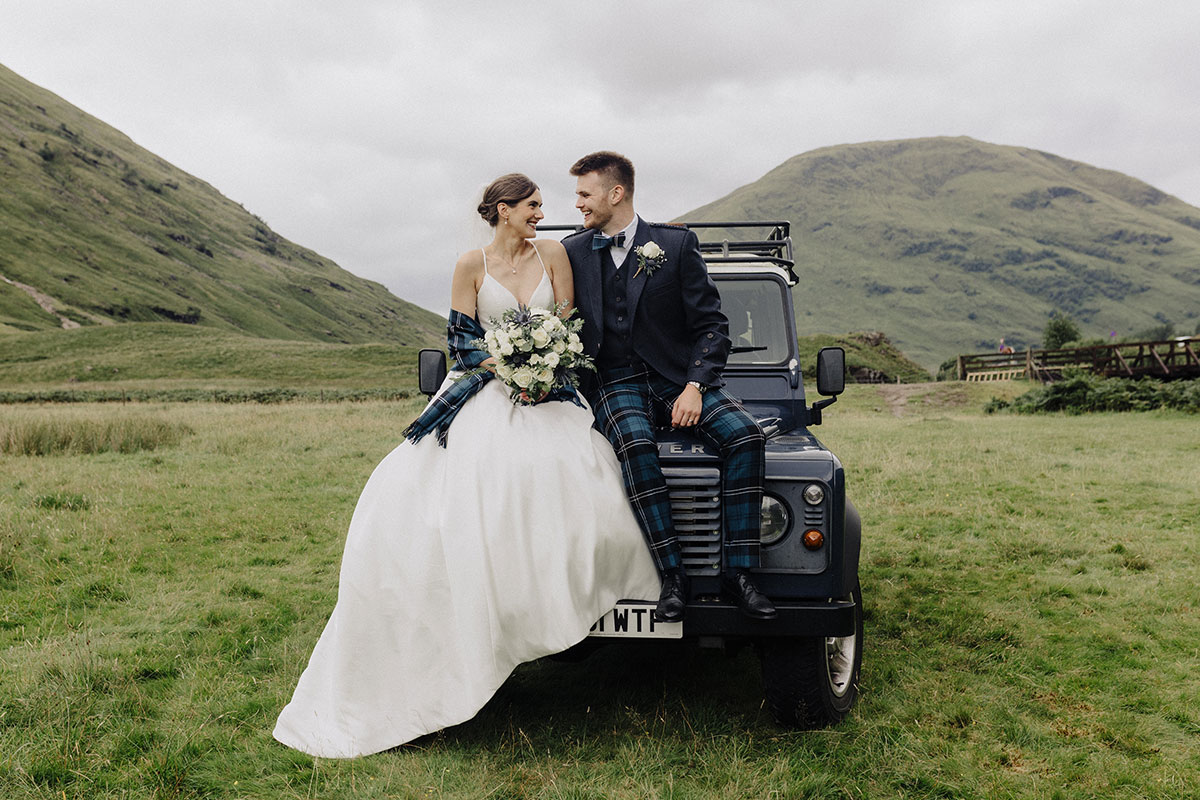 Bride and groom looking at one another, sitting on the hood of a land rover