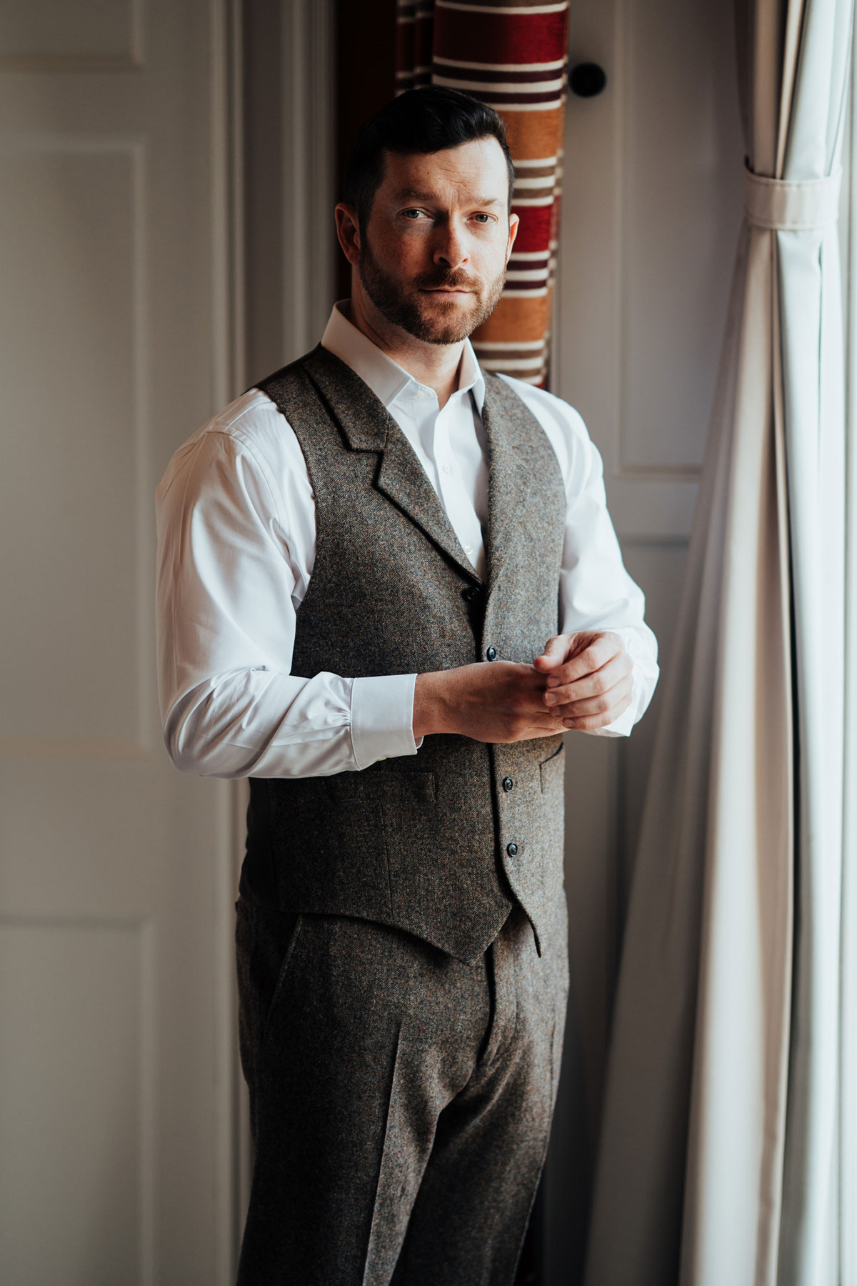 Groom in a brown tweed waistcoat fastening his cuffs beside a window at Schloss Roxburghe