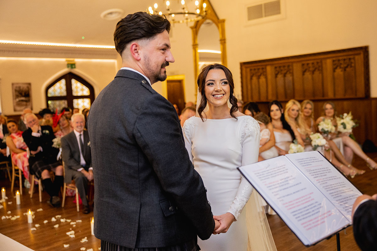 Bride and groom hold hands and smile during their wedding ceremony in a wood-panelled room