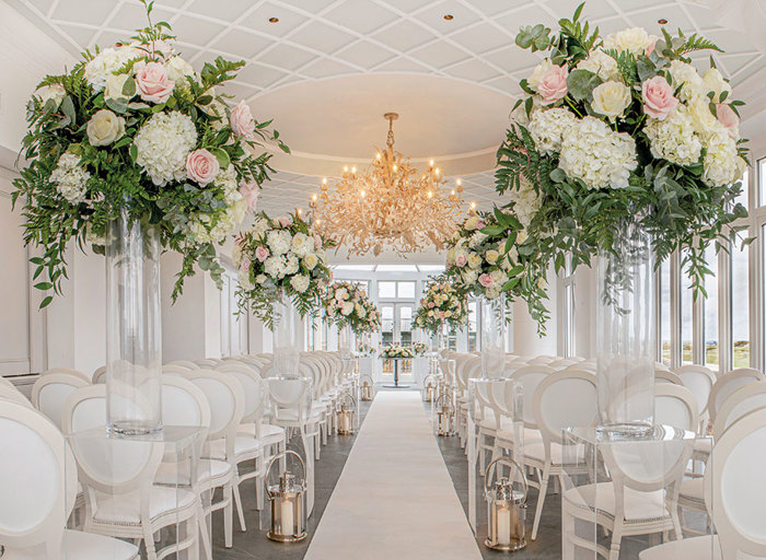 Rows of white chairs and white flower arrangements on either side of an aisle in a conservatory