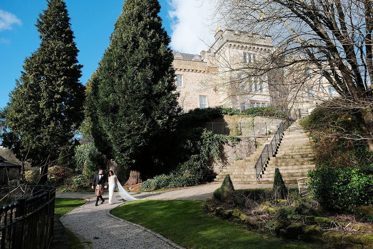 bride and groom walk hand in hand towards water with crossbasket castle in the background