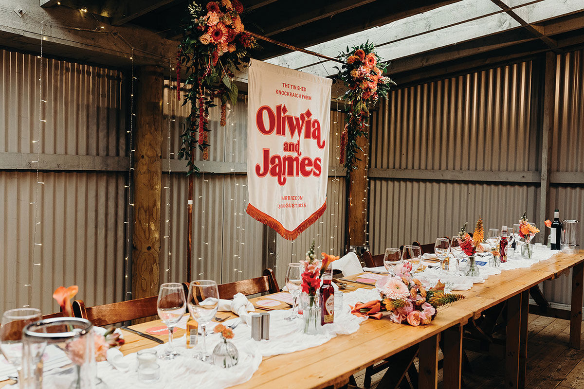 Rustic wedding reception table with vibrant floral arrangements, hanging installations and a personalised banner in a barn setting