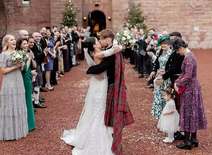 A couple kissing in front of a castle wearing a wedding dress and a red kilt with two rows of people standing behind them