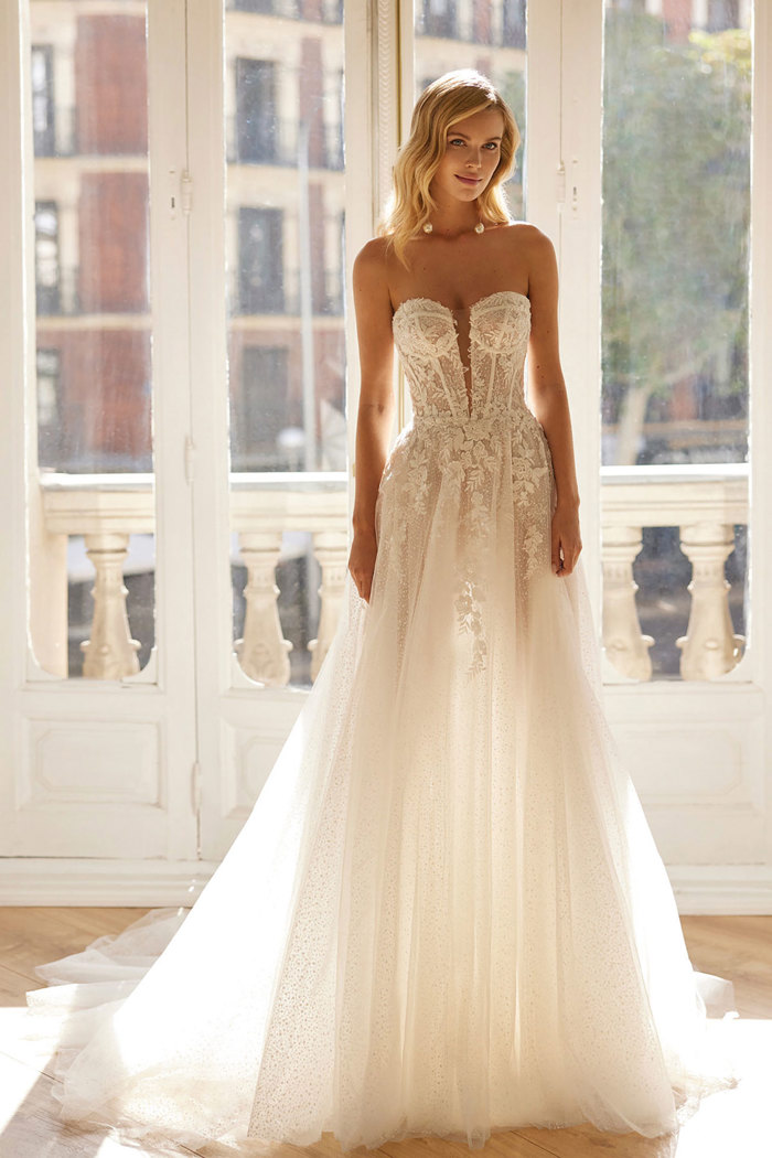 blonde bride stands with large window behind her, wearing a strapless v-neck ballgown with sheer tulle, a boned bodice and plenty of embellishment