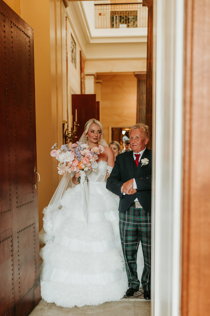 A bride wearing a wedding dress with a large skirt standing arm in arm with a man wearing tartan trousers and a suit jacket 