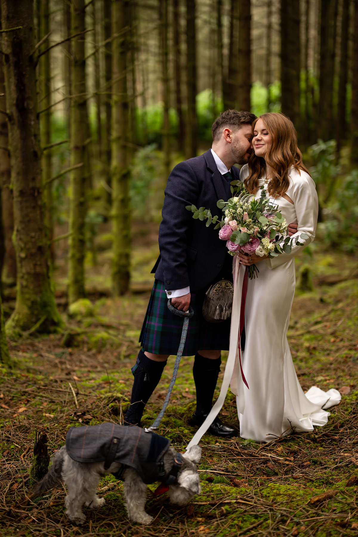 Bride and groom sharing a quiet moment in a Scottish woodland after their Glencoe elopement while their schnauzer explores nearby