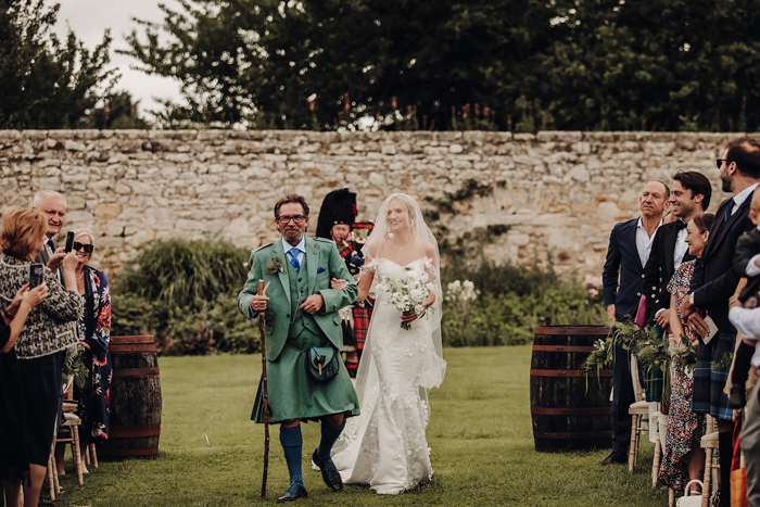 A Bride And Man In A Kilt Walking Down An Aisle Set Up In A Garden