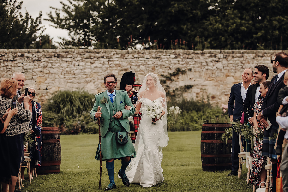 A Bride And Man In A Kilt Walking Down An Aisle Set Up In A Garden