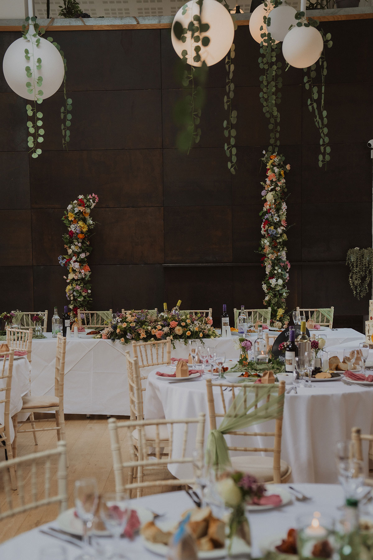 Floral-adorned top table with colourful arrangements and candlelight beneath hanging globe lights at Scottish wedding reception