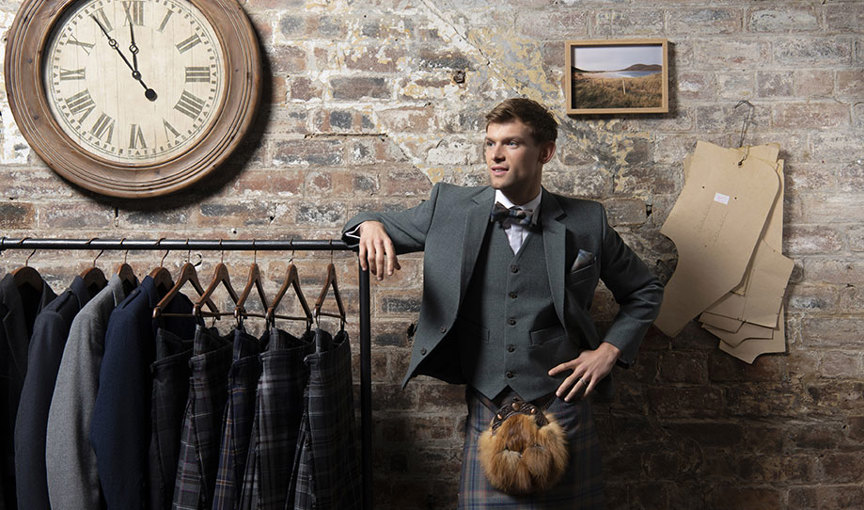 A person wearing a MacGregor and MacDuff Harris Tweed kilt outfit leaning on a clothes rail upon which jackets and kilts are hanging; rustic exposed brick wall in background with a card clothing pattern hanging on wall and large clock
