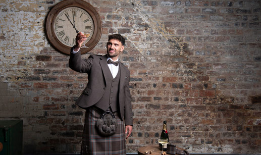 A person wearing a Jura Mist tartan kilt outfit from MacGregor and MacDuff raising a champagne glass. They are standing in front of a rustic brick wall underneath a large clock.