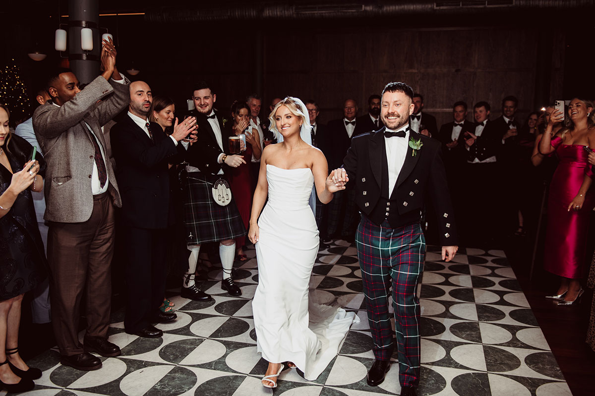 Bride and groom walking hand in hand across a black-and-white dancefloor as guests stand and applaud around them