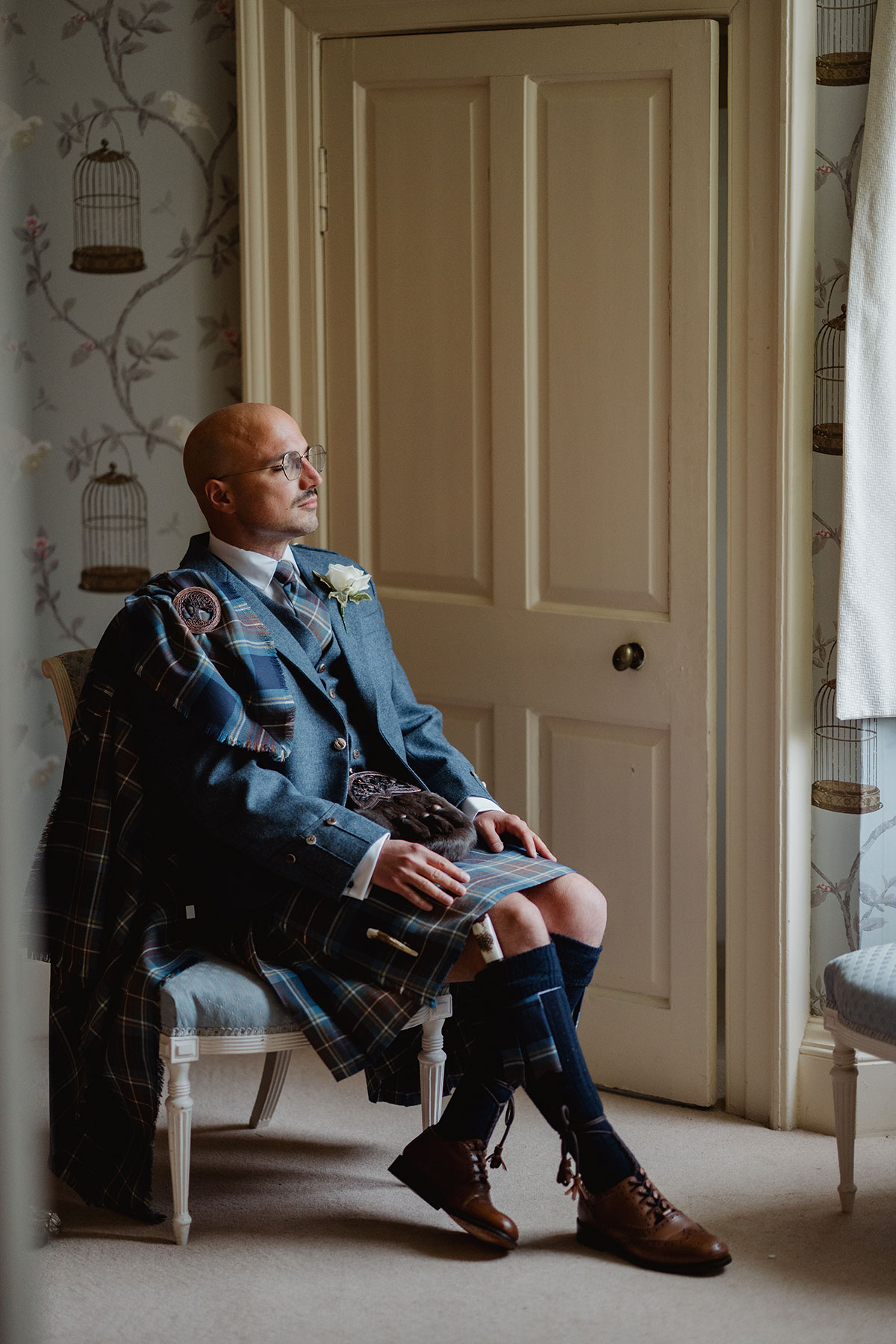 A groom in full Highland dress sits thoughtfully on a chair near a window, with soft light highlighting his tartan kilt and jacket