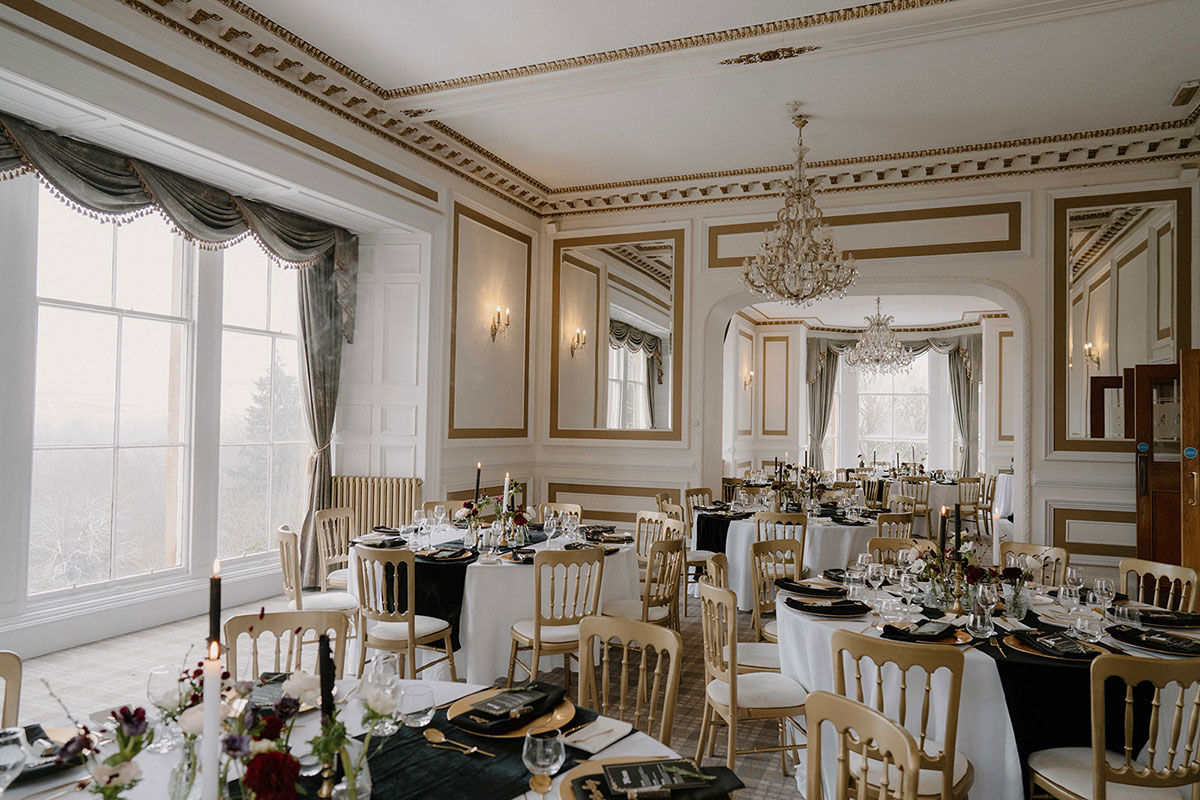 Wedding reception setup inside Auchen Castle with round dining tables, black and white place settings, chandeliers and large sash windows overlooking the grounds
