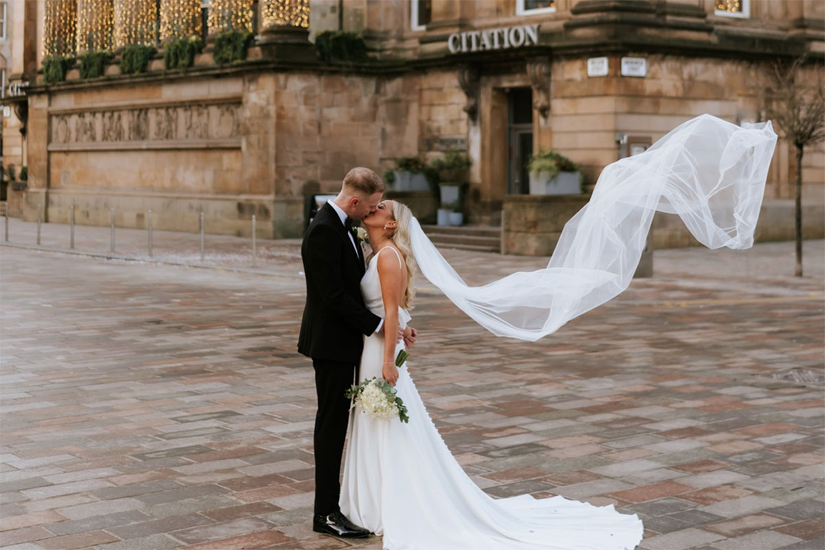 bride and groom kiss out on cobbled street with bride's veil floating in the wind and a columned. building labelled Citation directly behind them