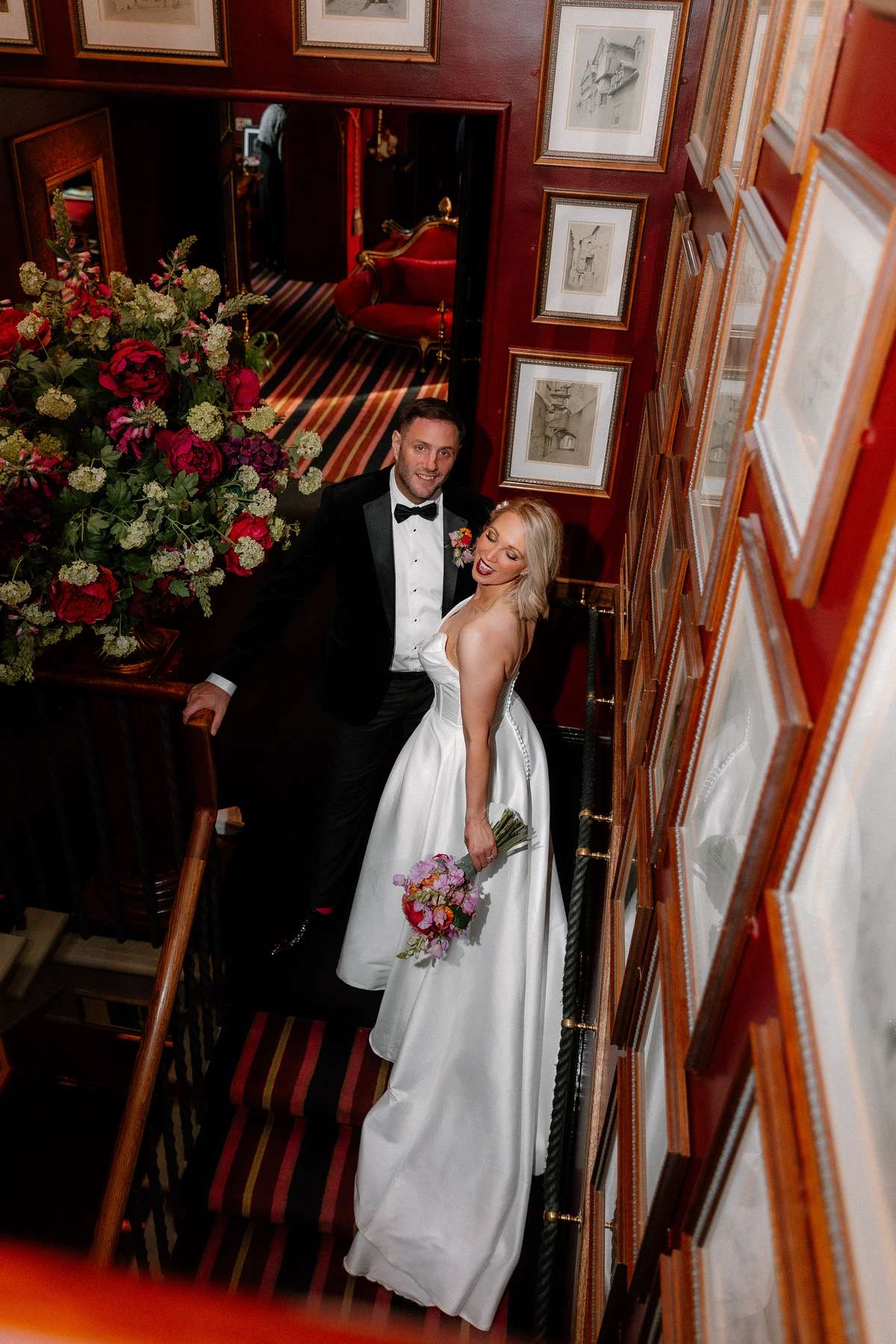 Kara and Rory pose together on the staircase inside Prestonfield House, surrounded by rich red interiors and floral displays.