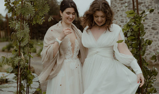 two laughing brides looking downward while standing outside under a foliage-covered wire arch in a garden with old stone building in background