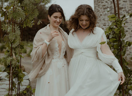 two laughing brides looking downward while standing outside under a foliage-covered wire arch in a garden with old stone building in background