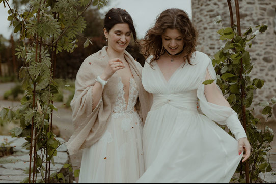 two laughing brides looking downward while standing outside under a foliage-covered wire arch in a garden with old stone building in background