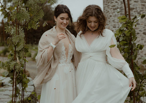 two laughing brides looking downward while standing outside under a foliage-covered wire arch in a garden with old stone building in background