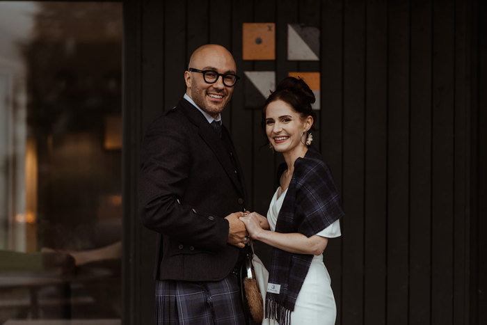 groom in blue, grey and white tartan kilt outfit holds hands with bride in white wedding dress with same tartan shawl over her shoulders