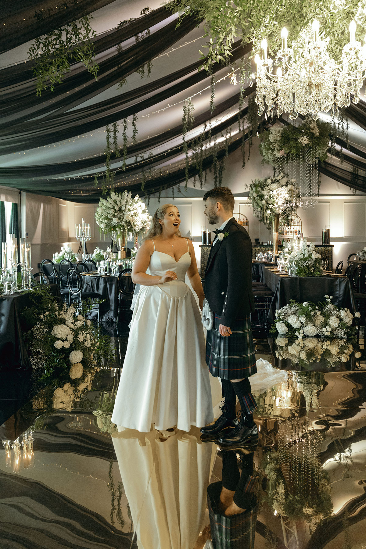 Bride in satin gown and groom in kilt share a moment beneath chandeliers at Dundas Castle’s romantasy-inspired reception.