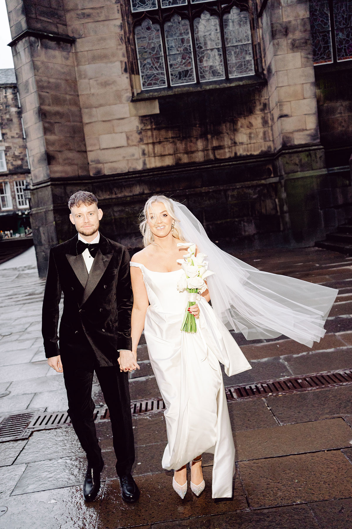 Newlyweds walking hand in hand outside a stone building in Edinburgh, with the bride’s veil flowing behind her as they smile towards the camera