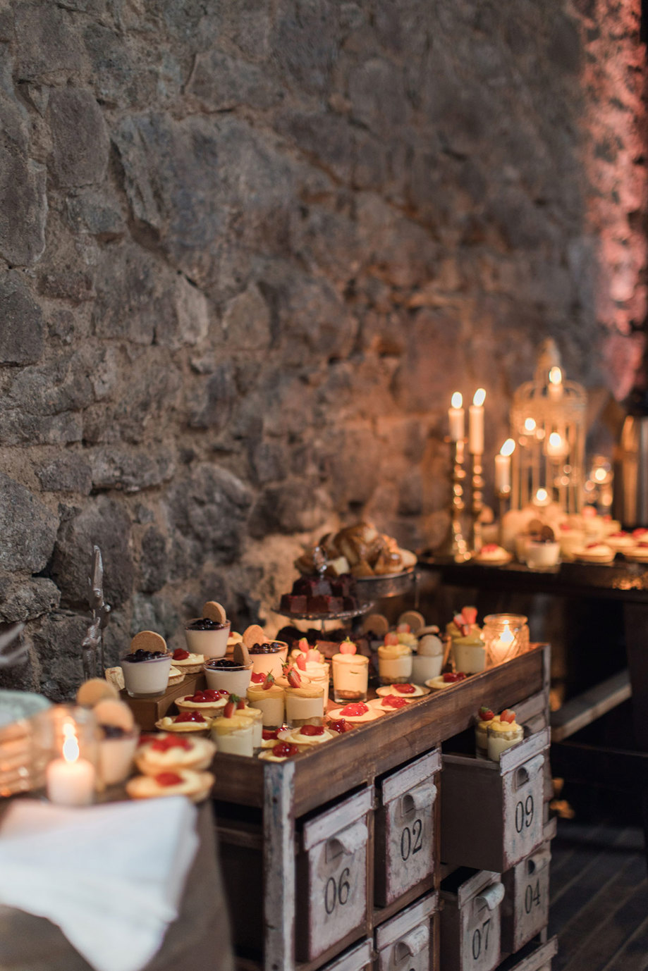 an old-looking wooden cabinet with numbered drawers with miniature cakes and desserts on top of it. It sits against a grey stone wall and there are candles flickering in the background.