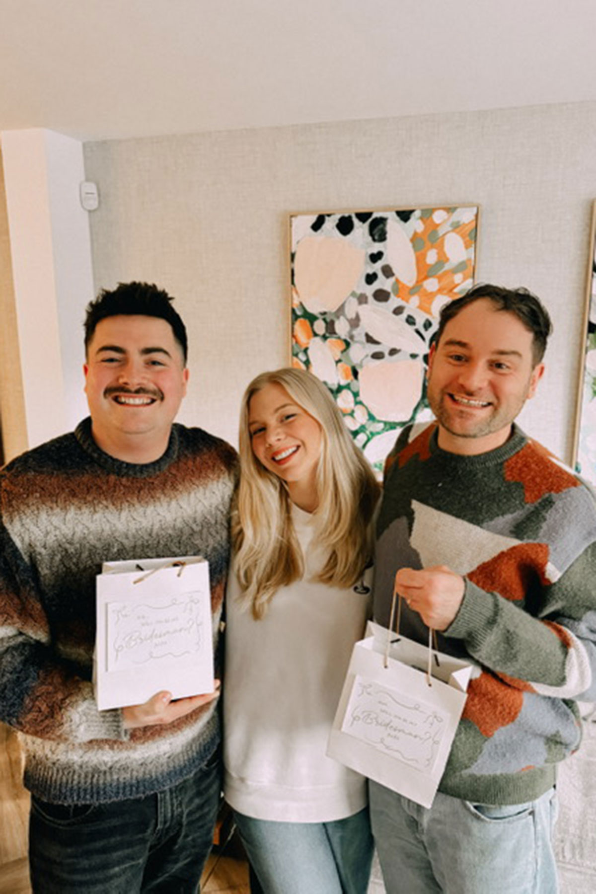Three friends smiling indoors holding gift bags in casual setting