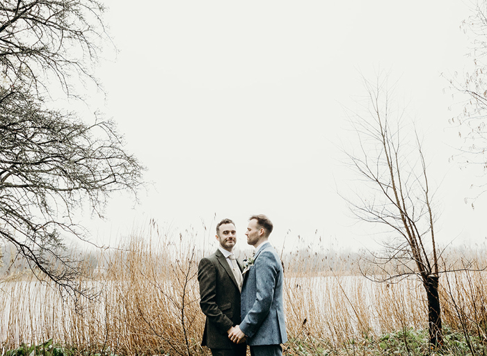 two grooms holding hands in front of tall golden grass and a body of water. The shot is framed by sparse-looking branches and tree