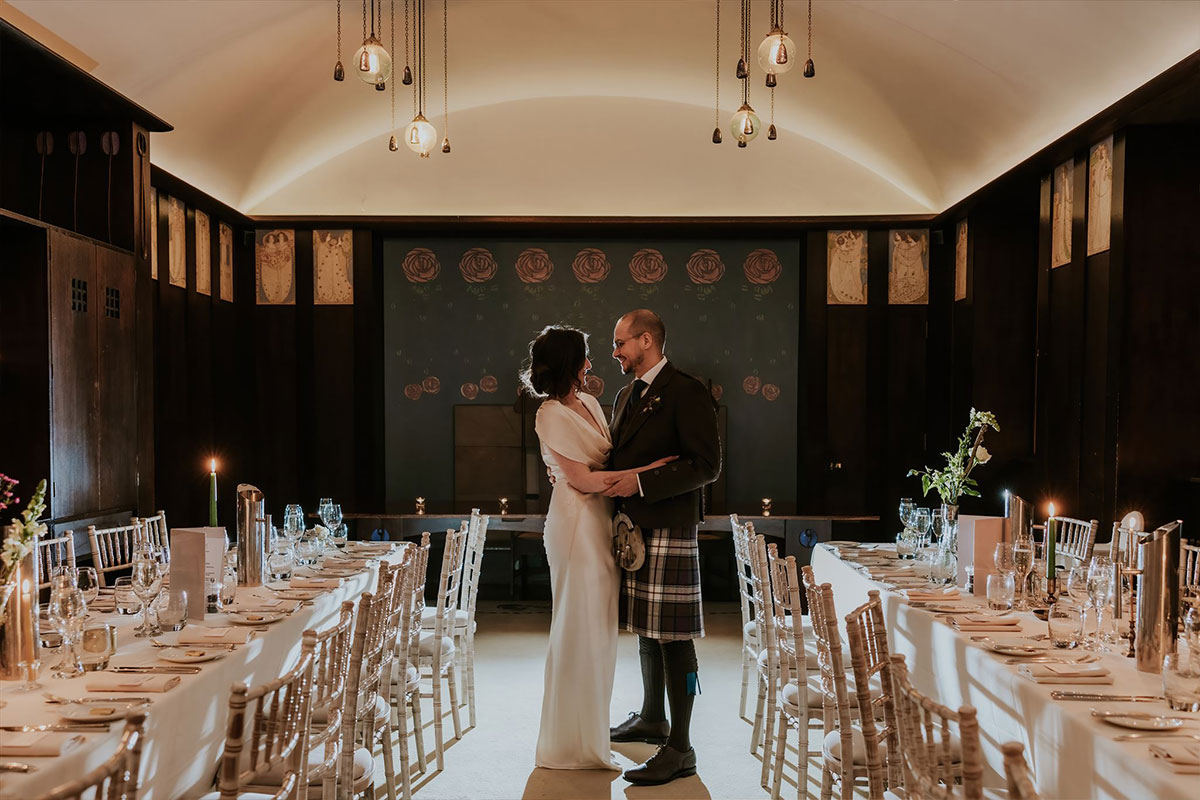 A bride in a white dress and a groom inn a kilt stand with their arms around each other next to two long dining tables