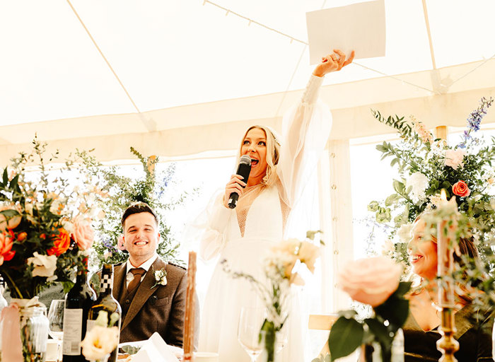 a bride holds a microphone and raises her arm in the air. The groom and another wedding guest sit either side of her at a table in a marquee. There are wine bottles and glasses on the table, floral decorations in the background