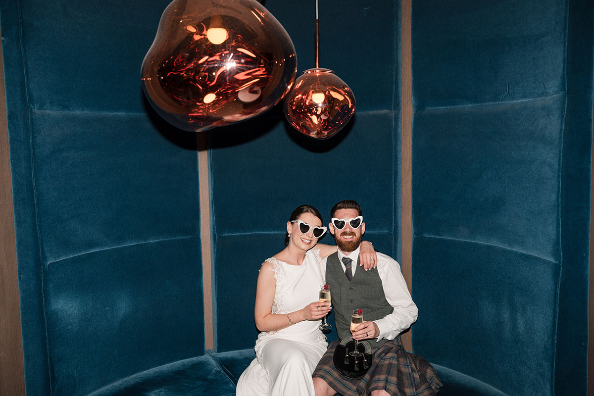 Bride and groom wearing heart-shaped sunglasses sit together with drinks at Clayton Hotel Glasgow wedding reception.
