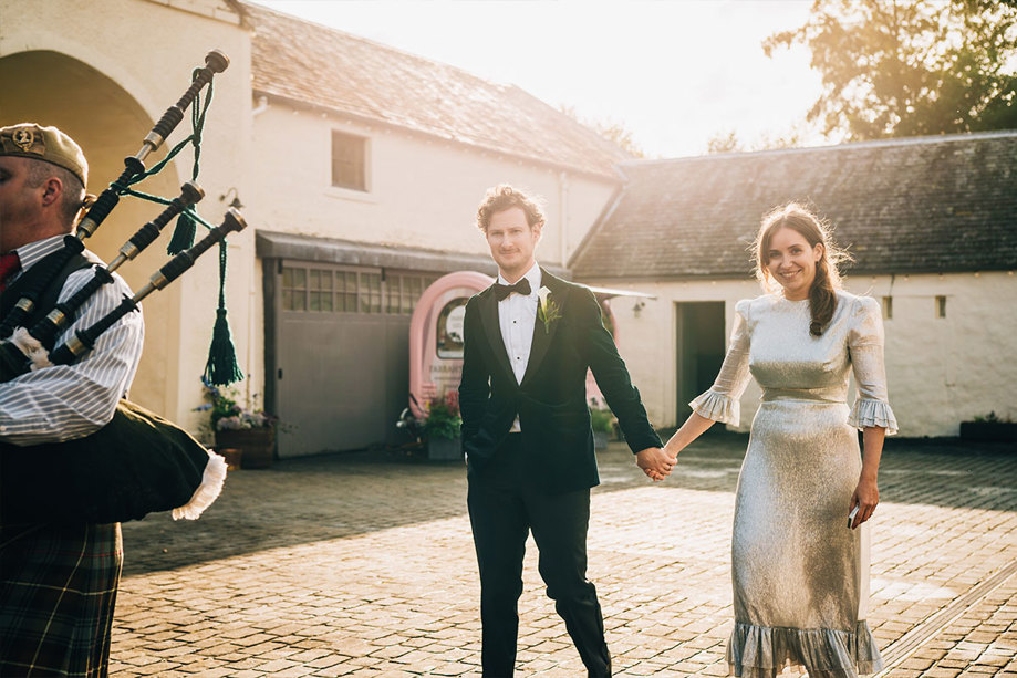 A bride and groom walking through a barn's courtyard following a bagpiper