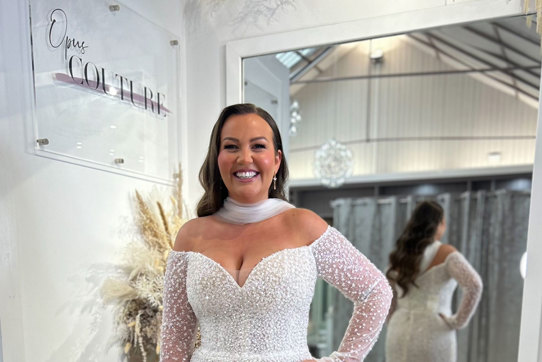 smiling dark-haired model wears off the shoulder long sleeve shimmery wedding dress and a tulle neck scarf